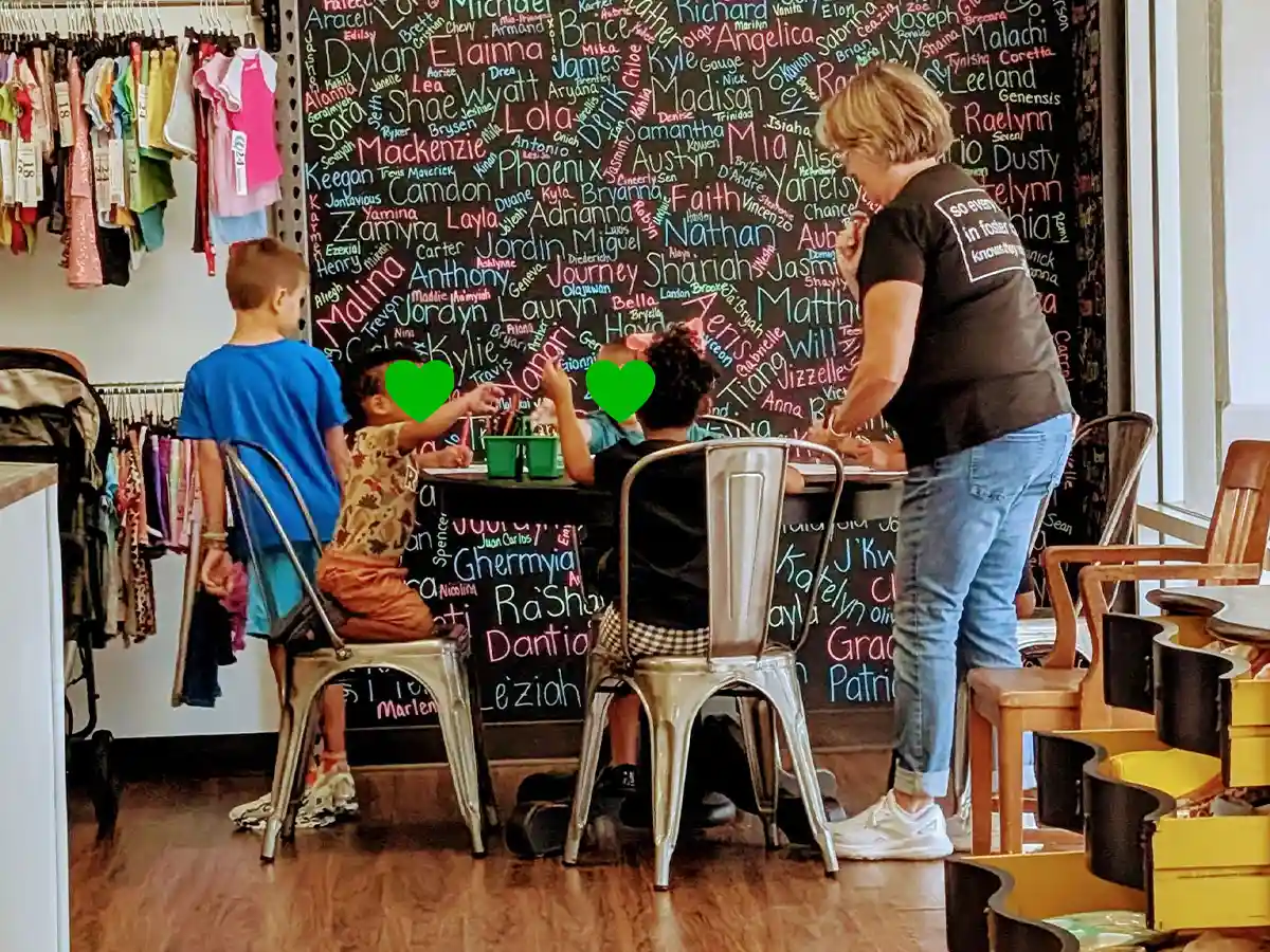 Children sitting at a table drawing with an adult helper in front of a name-filled chalkboard wall