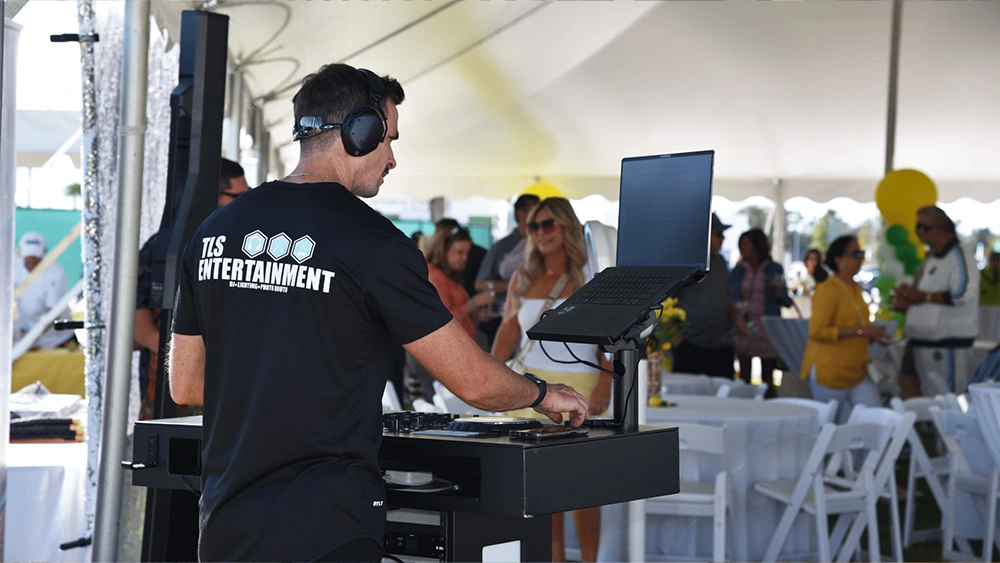 A DJ wearing a TLS Entertainment shirt mixing music on equipment under a festival tent.